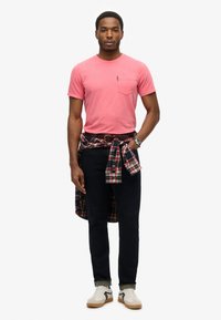 Camiseta corta de manga rosa con bolsillo, jeans oscuros y camisa a cuadros atada a la cintura. El calzado son zapatillas blancas con acentos negros.