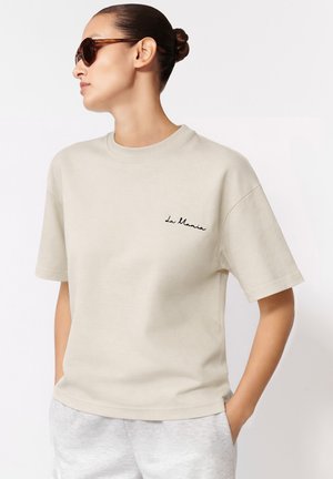 Kobieta ubrana w beżowy T-shirt z krótkim rękawem z napisem "La Mania" na piersi, okulary przeciwsłoneczne i jasnoszare spodnie, ręce w kieszeniach, patrząca na bok.