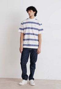 T-shirt bianca a maniche corte con righe blu e navy, con il logo "HUGO". Abbinata a jeans scuri in denim e sneakers bianche.