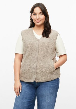 Frau mit dunklem Haar trägt eine beige strukturierte Weste über einem weißen Hemd und blauen Jeans, steht mit einer Hand in der Westentasche.