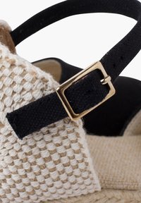 Beige woven sandal with black fabric strap and gold buckle detail. Features a textured surface and flat jute sole.
