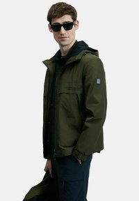 Veste imperméable vert olive avec une fermeture éclair à l'avant, des poignets réglables et deux poches avant. Portée sur un pull à capuche noir, le modèle tient un sac noir.