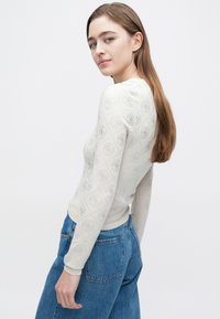 Maglione beige chiaro con motivo a diamante testurizzato e dettagli con logo, caratterizzato da maniche lunghe e design aderente. Abbinato a jeans blu.