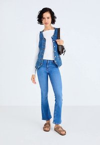 Traper prsluk s gumbima sprijeda, uparena s bijelom rebrastom majicom dugih rukava, svijetloplavim zvonolikim trapericama i smeđim sandalama.