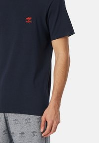Camiseta de algodón azul marino con mangas cortas, con un pequeño árbol de palma rojo bordado en el lado izquierdo del pecho. Diseño sencillo, corte clásico.