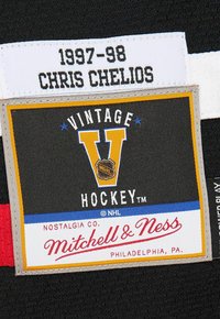 Étiquette de nom de Chris Chelios de 1997-98 au-dessus d'un écusson de hockey vintage Mitchell & Ness sur un tissu noir, faisant référence à la nostalgie des vêtements de la LNH.