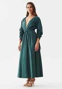 Teal pitkä mekko, jossa on syvä V-kaula-aukko, kerätty helma ja puffihihet. Sujuva kangas, jossa on nyörikiinnitys vyötäröllä. Beiget korolliset sandaalit.