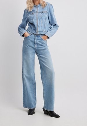 Kvinde iført lyseblå denimjakke og vidde jeans med sorte spidse ankelstøvler, stående med hænderne i lommerne.