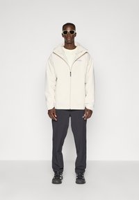 Light beige zip-up jacket with a hood, paired with dark gray pants and black athletic shoes. The jacket features a logo on the chest.