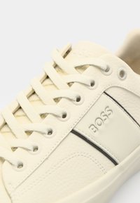 Baskets en cuir blanc avec surface texturée, lacets plats, détail du logo, et une fine bande noire sur le côté.