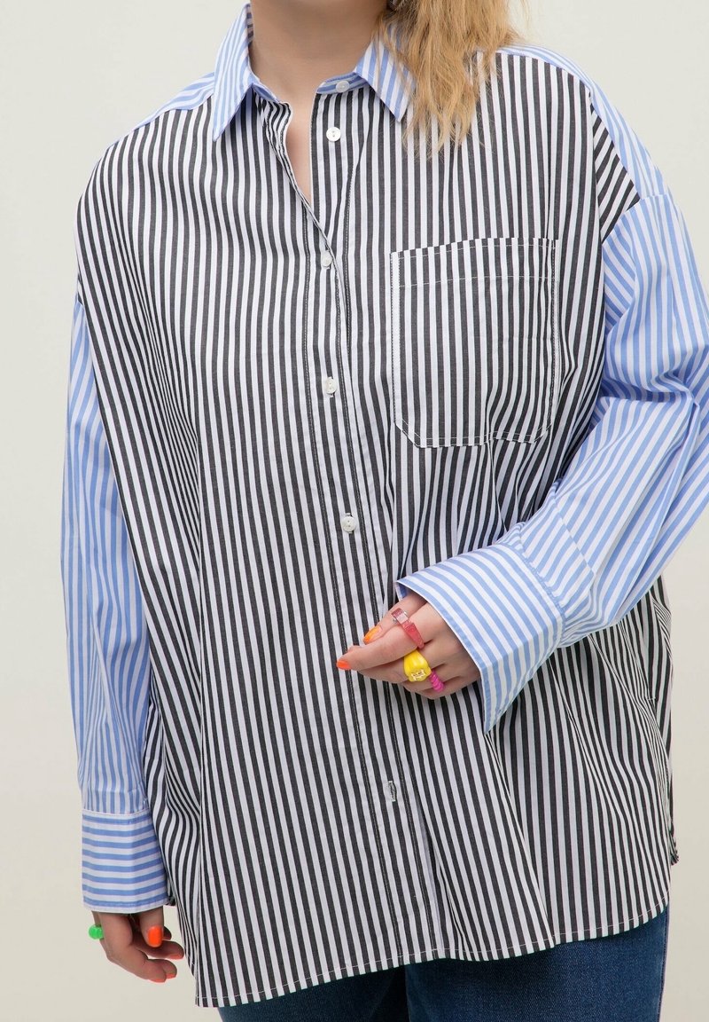 Chemise à manches longues avec des rayures verticales noires et blanches sur le devant et des manches à rayures verticales bleues et blanches, portée par une personne avec des bagues colorées.