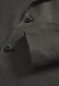 Tmavomodrý šedý materiál saka s jemnou diagonálnou textúrou. Obsahuje čierne gombíky so štyrmi dierkami a zložený manžetový detail.