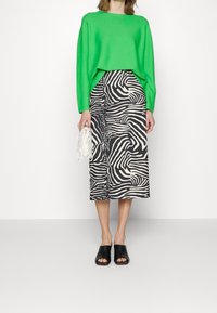 Pull vert, jupe midi à motif zèbre noir et blanc, mules noires à bout ouvert, tenant un petit sac à main blanc. Textures lisses.