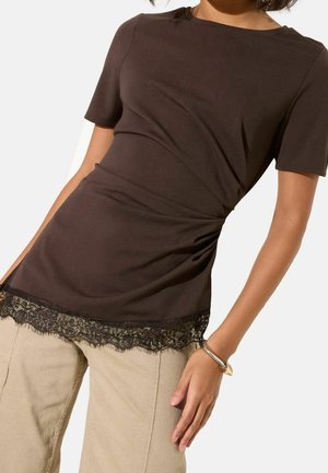 Femme portant un haut ajusté à manches courtes marron foncé avec une bordure en dentelle à l’ourlet et un pantalon beige clair, debout avec un bras détendu.