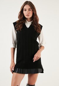 Black knitted sleeveless dress with a v-neck, ribbed texture, and pleated black skirt. Worn over a white collared shirt.
