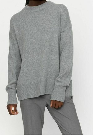 Personne portant un pull ample en maille gris clair et un pantalon gris assorti, debout devant un fond blanc uni.