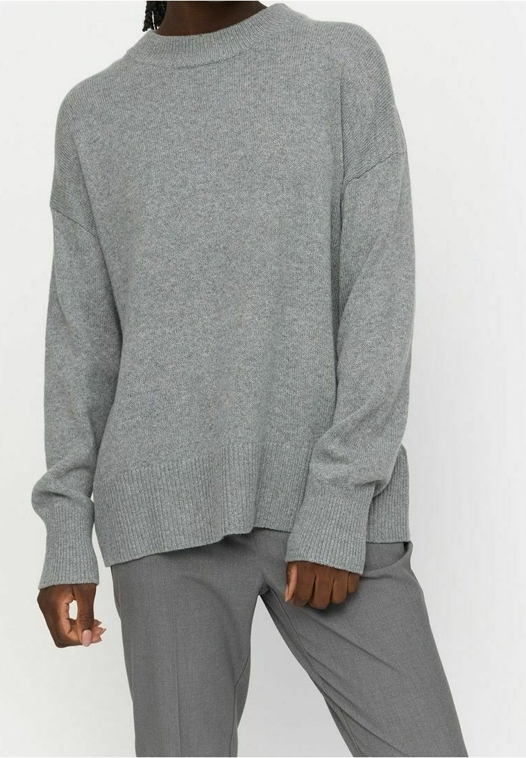 Personne portant un pull ample en maille gris clair et un pantalon gris assorti, debout devant un fond blanc uni.