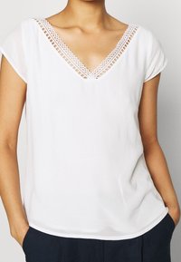 Blouse blanche à manches courtes avec un décolleté en V, ornée d'une dentelle décorative le long de l'encolure et en tissu lisse et léger.