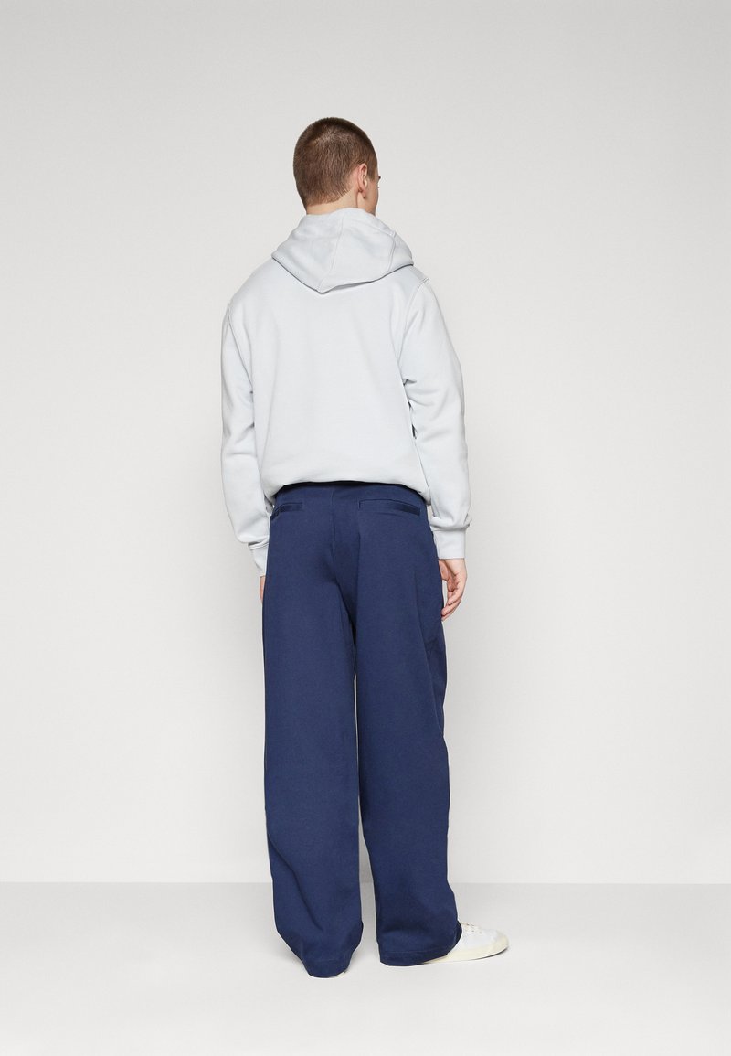 Uomo di spalle che indossa un felpa grigia chiara, pantaloni larghi blu navy e sneakers bianche su uno sfondo chiaro e uniforme.