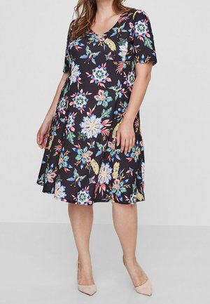 Femme portant une robe noire mi-longue ornée de motifs floraux colorés et à manches courtes, associée à des escarpins nus à bout pointu.