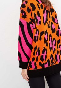 Pull vif avec des motifs animaliers orange vif et rose, porté par une personne vue de dos et de côté, associé à un pantalon noir.