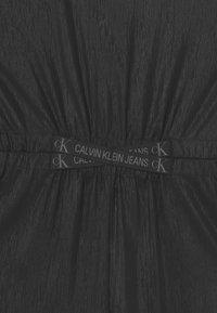 Czarna tkanina o teksturowanym wykończeniu, z marszczonym pasem i wyraźnym pasem oznaczonym napisem "CALVIN KLEIN JEANS" w odcieniach szarości.