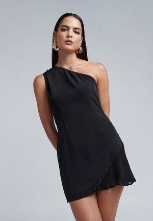 Sort étuikjole med én skulder, tætsiddende bodice, plisseret asymmetrisk bund, glat stofstruktur og slank silhuet. Guldøreringe fremhæver.