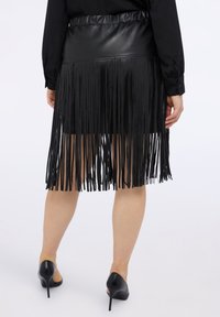 Czarna spódnica z sztucznej skóry z frędzlami, teksturowanym pasem i dopasowanym krojem. Nosi ją osoba w czarnych szpilkach.