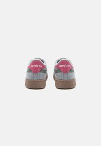 Adidași din suede gri cu accente roz, având o talpă din cauciuc texturat maro și un guler căptușit. Logo-ul "Reebok" pe toc.