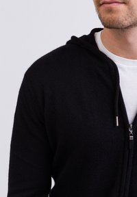 Felpa nera con zip realizzata in morbido tessuto texturizzato, dotata di cappuccio e zip in metallo argentato, indossata sopra una maglietta bianca.