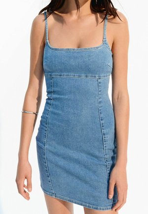 Femme portant une mini robe en jean bleu à coupe ajustée avec de fines bretelles et un bracelet argenté sur le bras gauche, sur fond blanc.