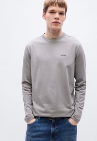 Γκρι μακρυμάνικο πουκάμισο από λείο βαμβακερό μείγμα, με στρογγυλή λαιμόκοψη και μικρό λογότυπο «BOSS» στο στήθος. Φορεμένο με τζιν παντελόνι.