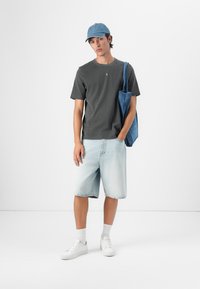 Grauer Baumwoll-T-Shirt, hellblaue Jeansshorts, weiße Sneaker und eine blaue Cap. Das Outfit ist lässig, mit einer Jeans-Tote-Tasche über einer Schulter getragen.