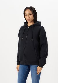 Cazaco preto com fecho éclair, capuz forrado, grandes bolsos e punhos canelados. Feito de um tecido macio com um ajuste descontraído, adequado para uso casual.