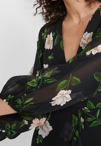 Zwarte bloemenblouse met doorzichtig materiaal, V-hals en gepofte mouwen met geplooide manchetten. Witte en groene bloemenpatroon overal.