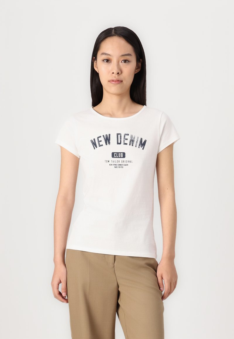 T-shirt bianca in cotone con maniche corte e colletti rotondo. Presenta una stampa grafica blu con la scritta "NEW DENIM CLUB". Indossata con pantaloni beige.