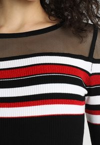 Sweter w paski w kolorach czarnym, białym i czerwonym, o ribbowanej fakturze. Przezroczysta czarna siateczka w dekolcie dodaje kontrastu i detali.