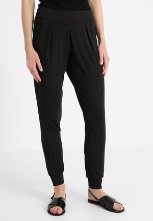 Vrouw draagt zwarte taps toelopende joggerbroek met zijzakken en omgeslagen enkels, gecombineerd met zwarte sandalen met open tenen, staand op een witte vloer.