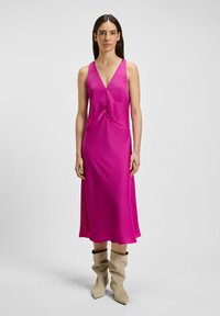 Fuchsia satijnen maxi-jurk met een diepe V-hals, gerimpeld detail bij de taille en een vloeiende silhouet; gecombineerd met beige slouchy laarzen.