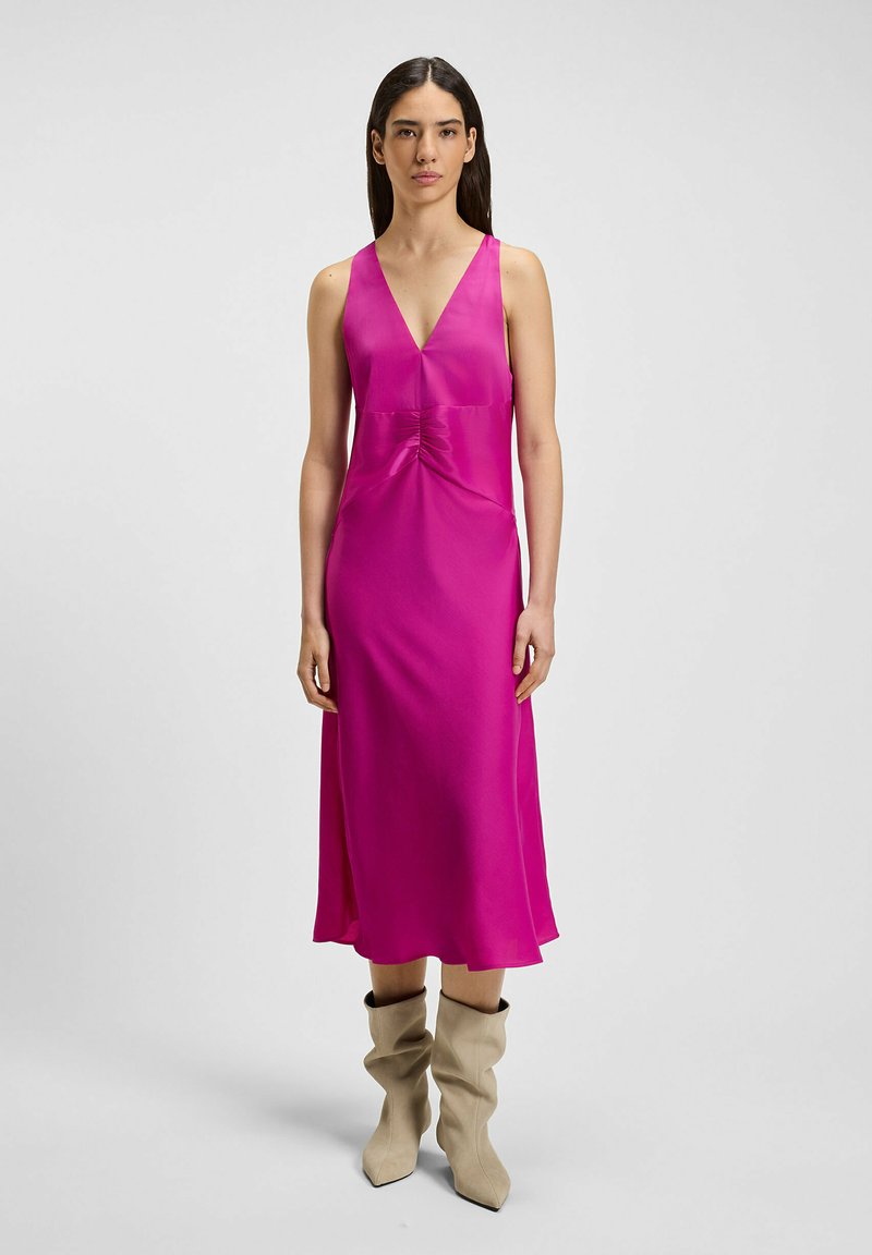Fuchsia satijnen maxi-jurk met een diepe V-hals, gerimpeld detail bij de taille en een vloeiende silhouet; gecombineerd met beige slouchy laarzen.