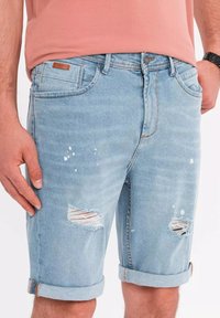 Ljusblå denimshorts har slitna detaljer med fransade hål, uppvikta ärmar och subtila färgstänk.