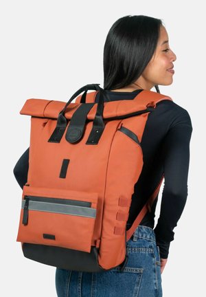 Oranger Rolltop-Rucksack mit schwarzen Trägern, einer grauen Reißverschlusstasche und einer strukturierten Oberfläche, der ein rundes Logo und seitliche Riemen aufweist.
