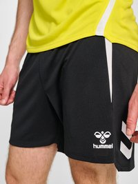 Hombre vestido con pantalones cortos deportivos negros Hummel con rayas blancas y camisa deportiva amarilla, de pie con las manos relajadas a los lados.