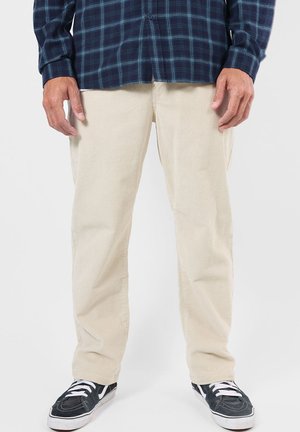 Homme portant un pantalon beige en velours côtelé, des baskets noires et blanches, et une chemise à carreaux bleu foncé, debout devant un fond uni.