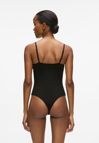 Juodas bodis su plonais reguliuojamais dirželiais, be siūlių dizainu ir prigludusia forma su aukštu nugarkaulio kirpimu. Lygus audinio paviršius.