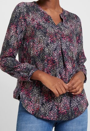 Femme portant un chemisier à manches longues avec un motif abstrait de points multicolores en bleu marine, rose, blanc et vert, associé à un jean bleu.