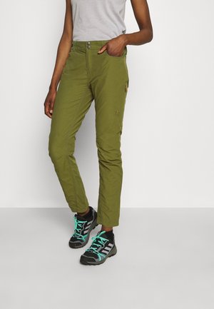 Person wearing olive green pants and gray athletic shoes with teal laces, standing with one hand in pocket against white background.