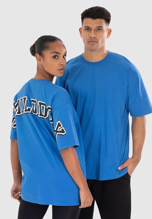 Blauwe katoenen t-shirts met korte mouwen. De ene heeft zwarte tekst op de achterkant, terwijl de andere eenvoudig is. Beide hebben een losse pasvorm.