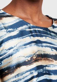 Blå och beige topp med rundad halsringning, abstrakta vågiga ränder och en strukturerad tyg, med synliga sömdetaljer.