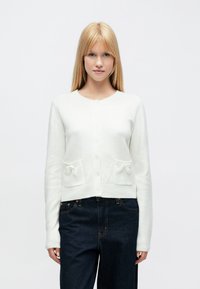 Fehér kötött kardigán hosszú ujjakkal, kerek nyakkal és két első zsebbel, amelyeken masni díszítések találhatók. Puha tapintású és rövidített fazonú.